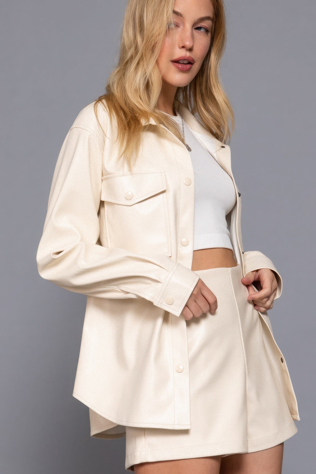 Woman wearing a chic, cream faux leather shacket over a matching mini skirt and crop top, perfect for stylish layering.