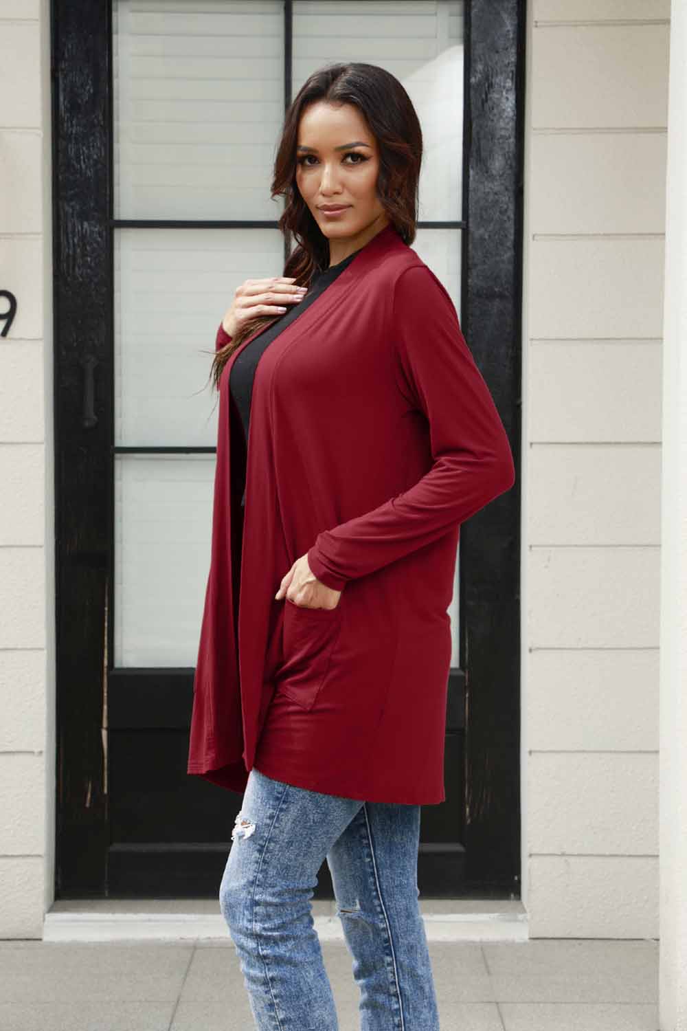 Woman wearing a burgundy open front long sleeve cardigan with pockets, paired with jeans, standing in front of a door.