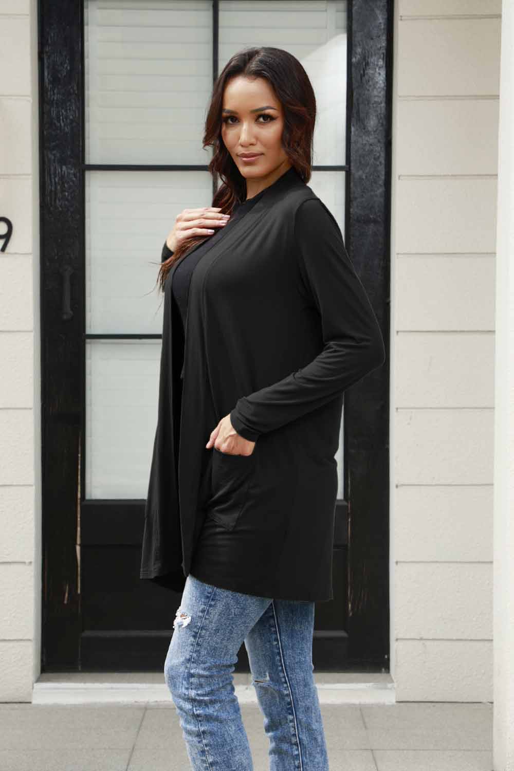 Woman wearing a black open front long sleeve cardigan with pockets, styled with jeans, standing in front of a door.