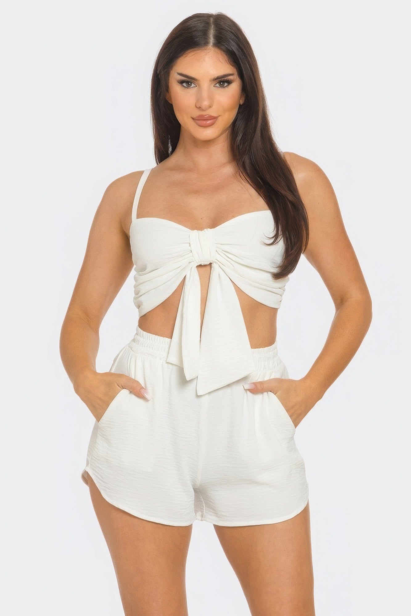 Woman wearing chic white shorts set with oversized front bow and twisted tie back, featuring elastic waistband and side pockets.