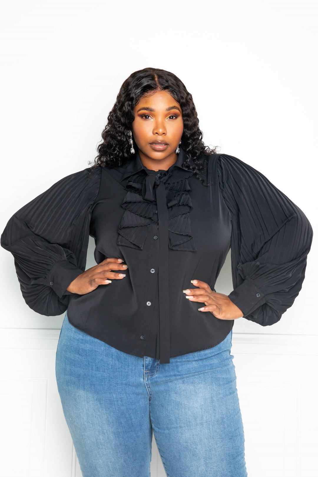 Plus-size model wearing a black pleated sleeve blouse with waterfall frill and bow, paired with blue jeans.