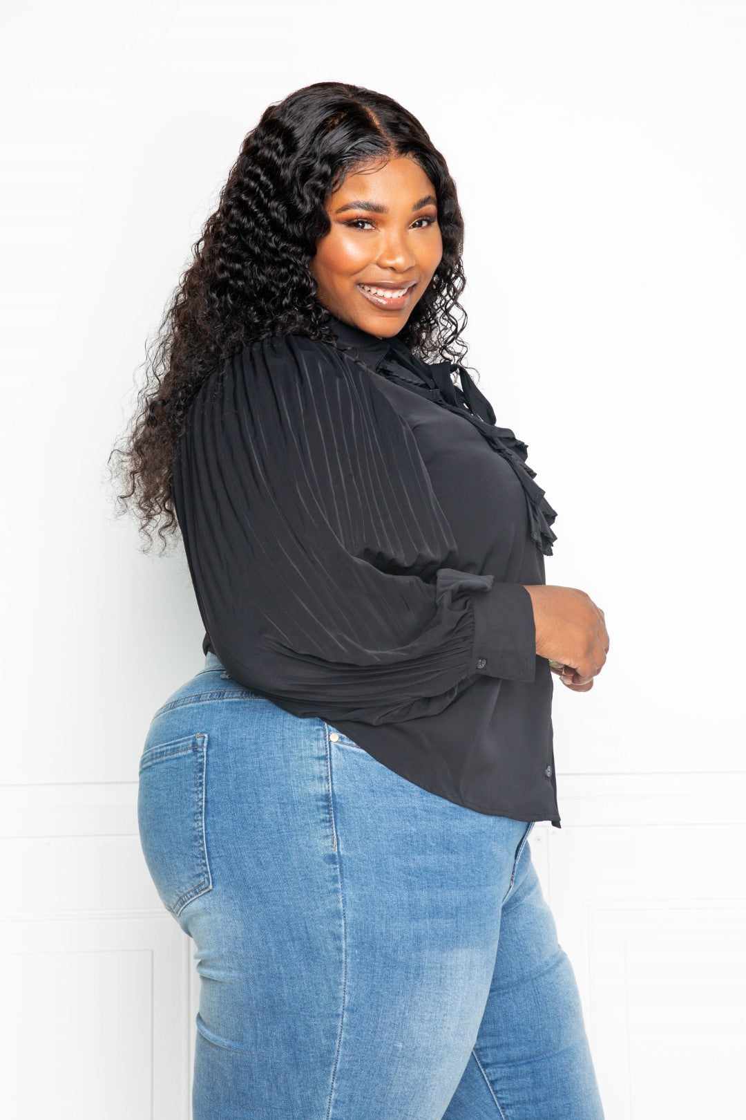 Plus size model in black pleated sleeve blouse with bow, featuring waterfall frill, paired with jeans. Perfect for stylish and elegant looks.