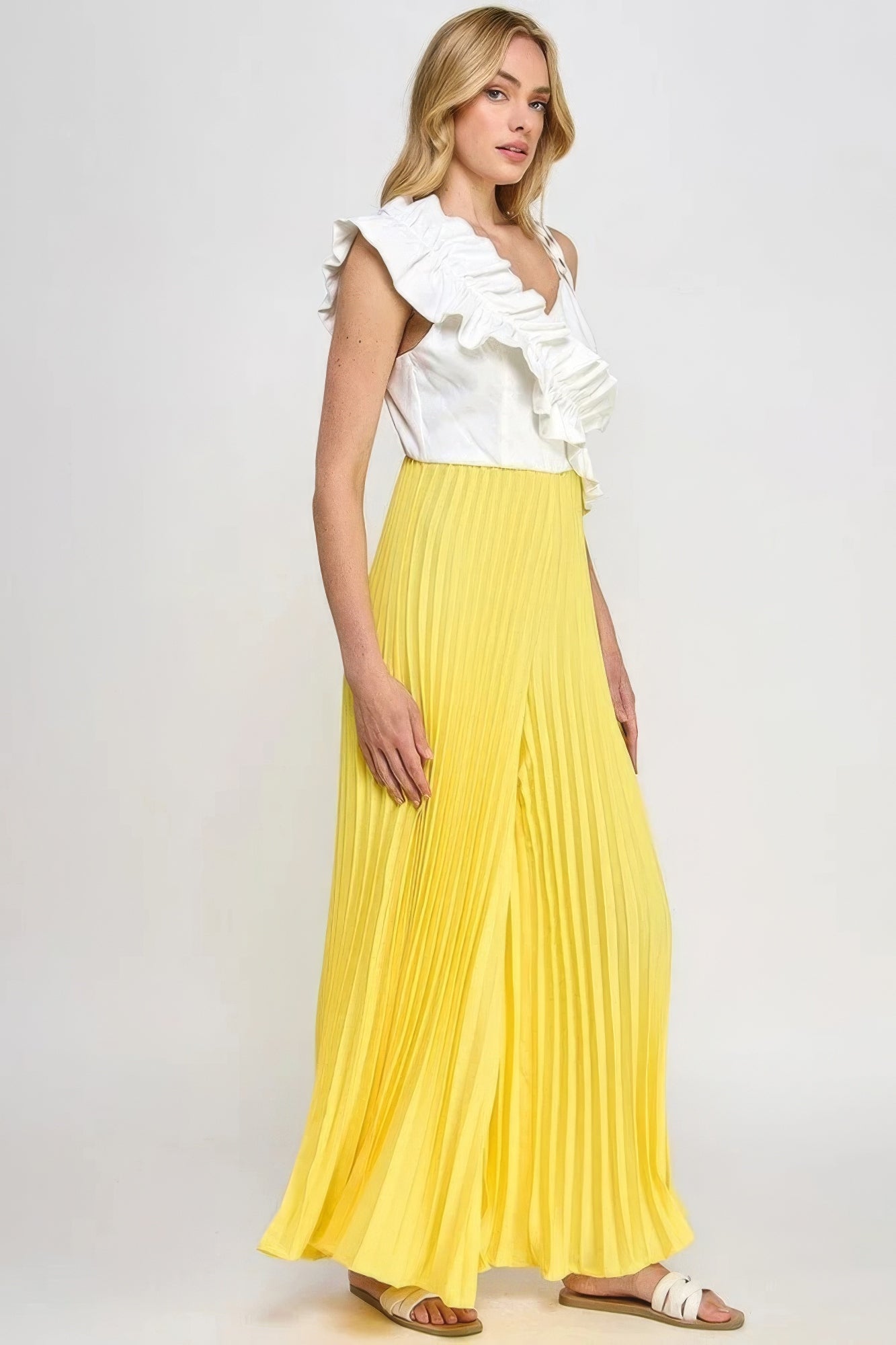 Woman wearing chic pleated cami jumpsuit with ruffle detailing in yellow and white, showcasing contemporary style and asymmetrical design.