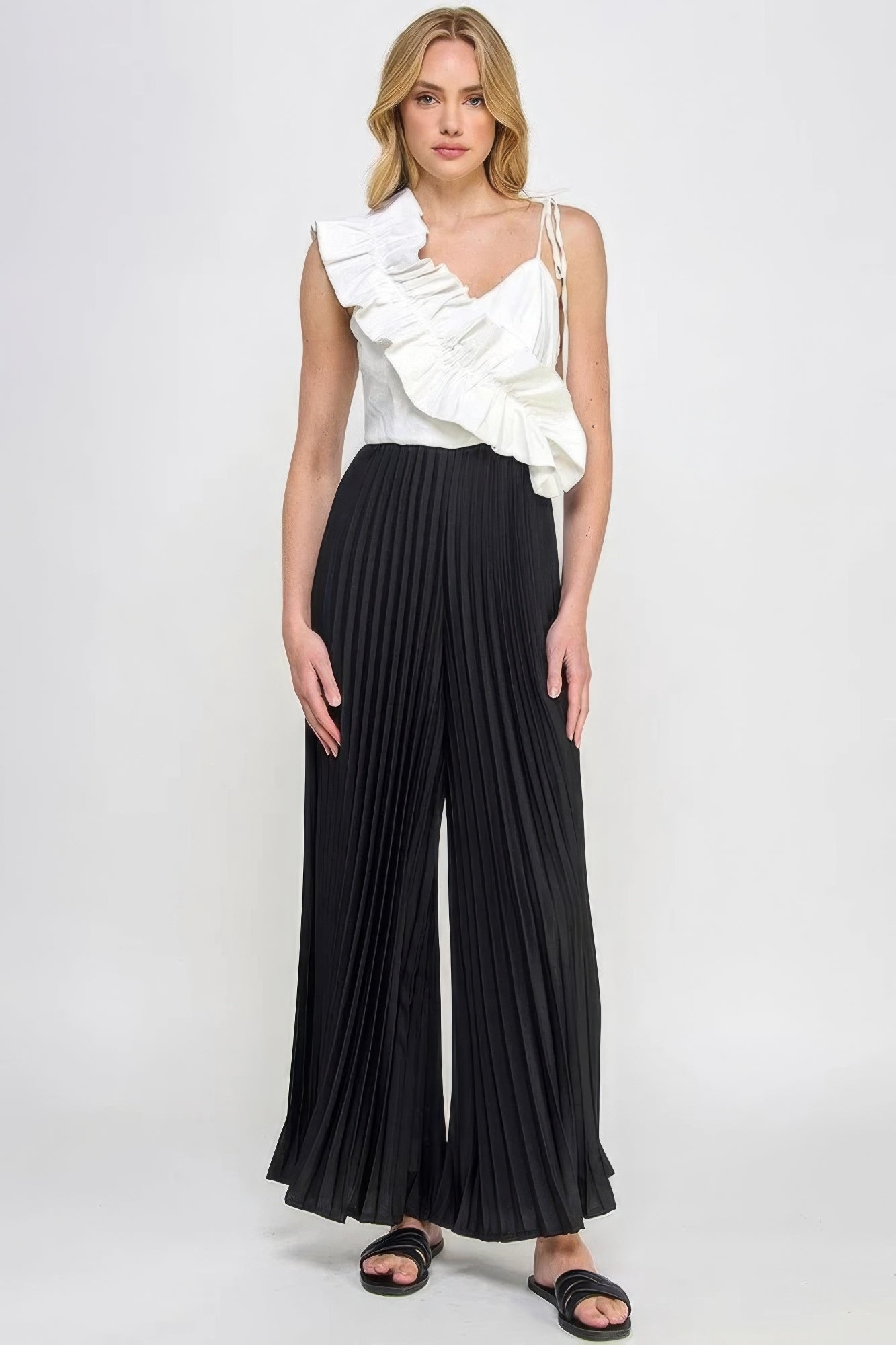 Woman wearing chic pleated cami jumpsuit with asymmetrical ruffle detailing and cami-style top, in black and white.