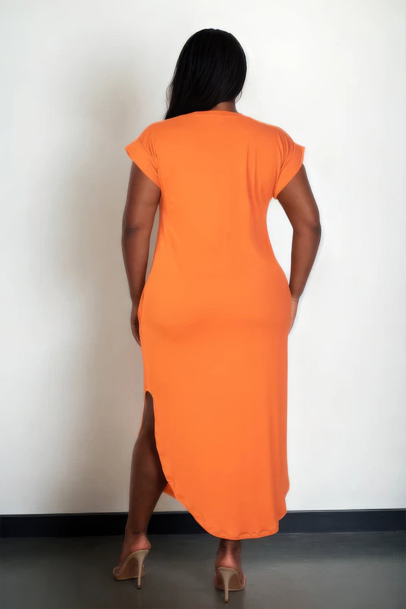 Woman wearing orange plus size maxi dress with side slit, viewed from the back, standing against a plain background.