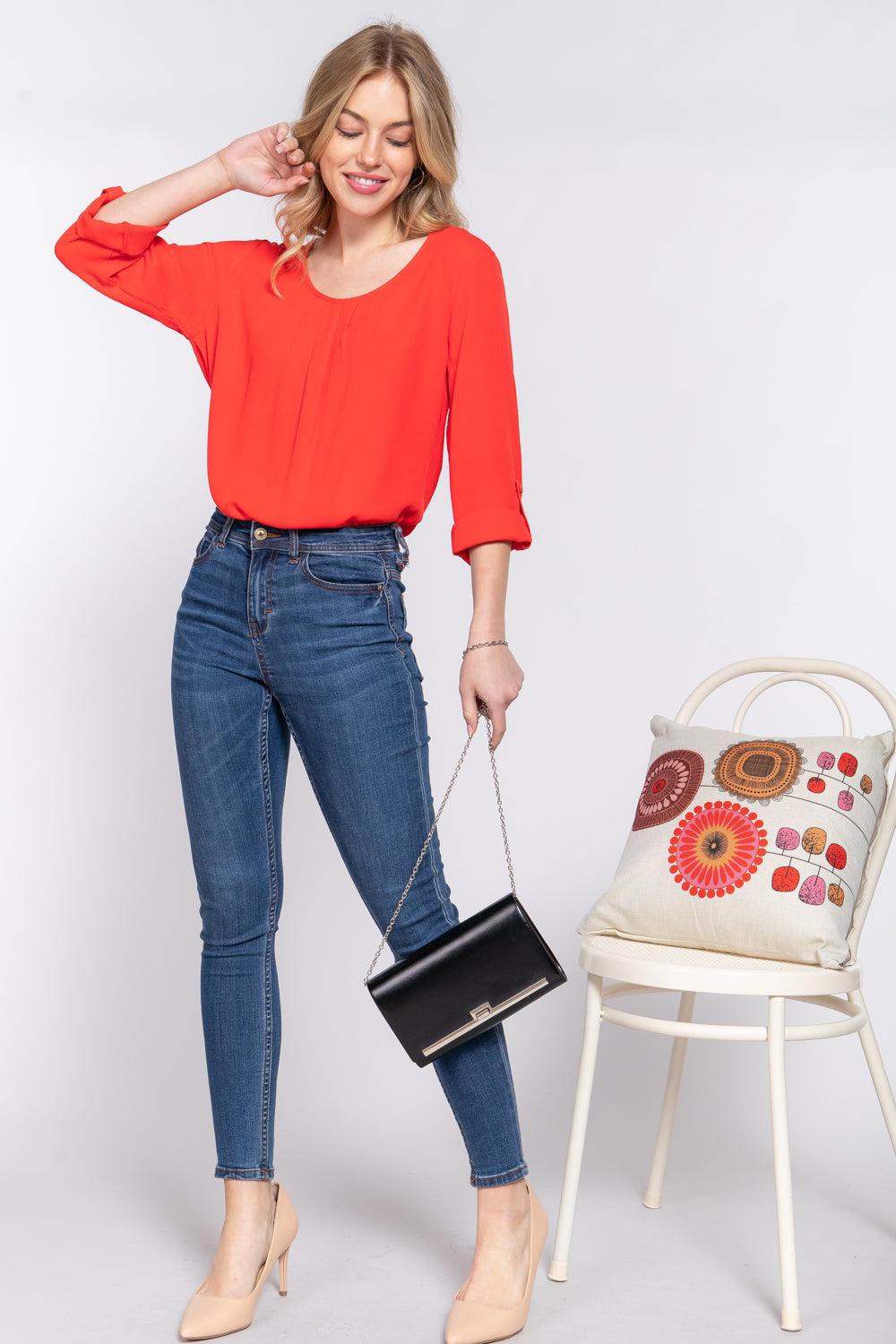 Woman wearing tomato red roll up sleeve blouse with pleats, paired with jeans, holding a black purse and standing beside a decorative chair.