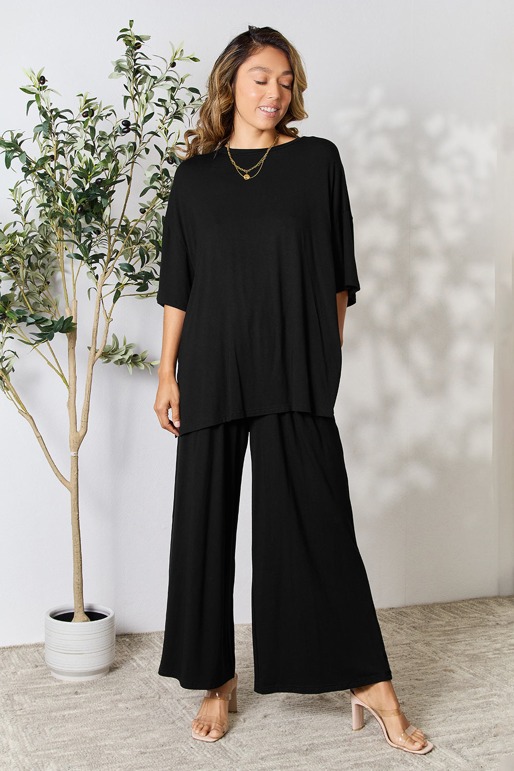 Woman wearing black round neck slit top and pants set in a stylish and comfortable design, standing next to a potted plant.