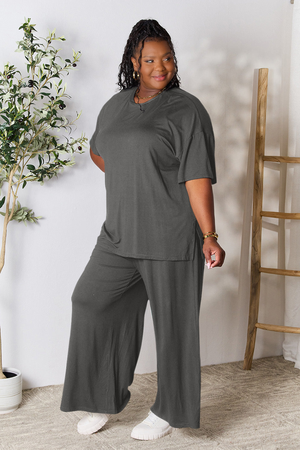 Woman wearing Double Take Round Neck Slit Top and Pants Set in dark gray, standing in a stylish room setting.