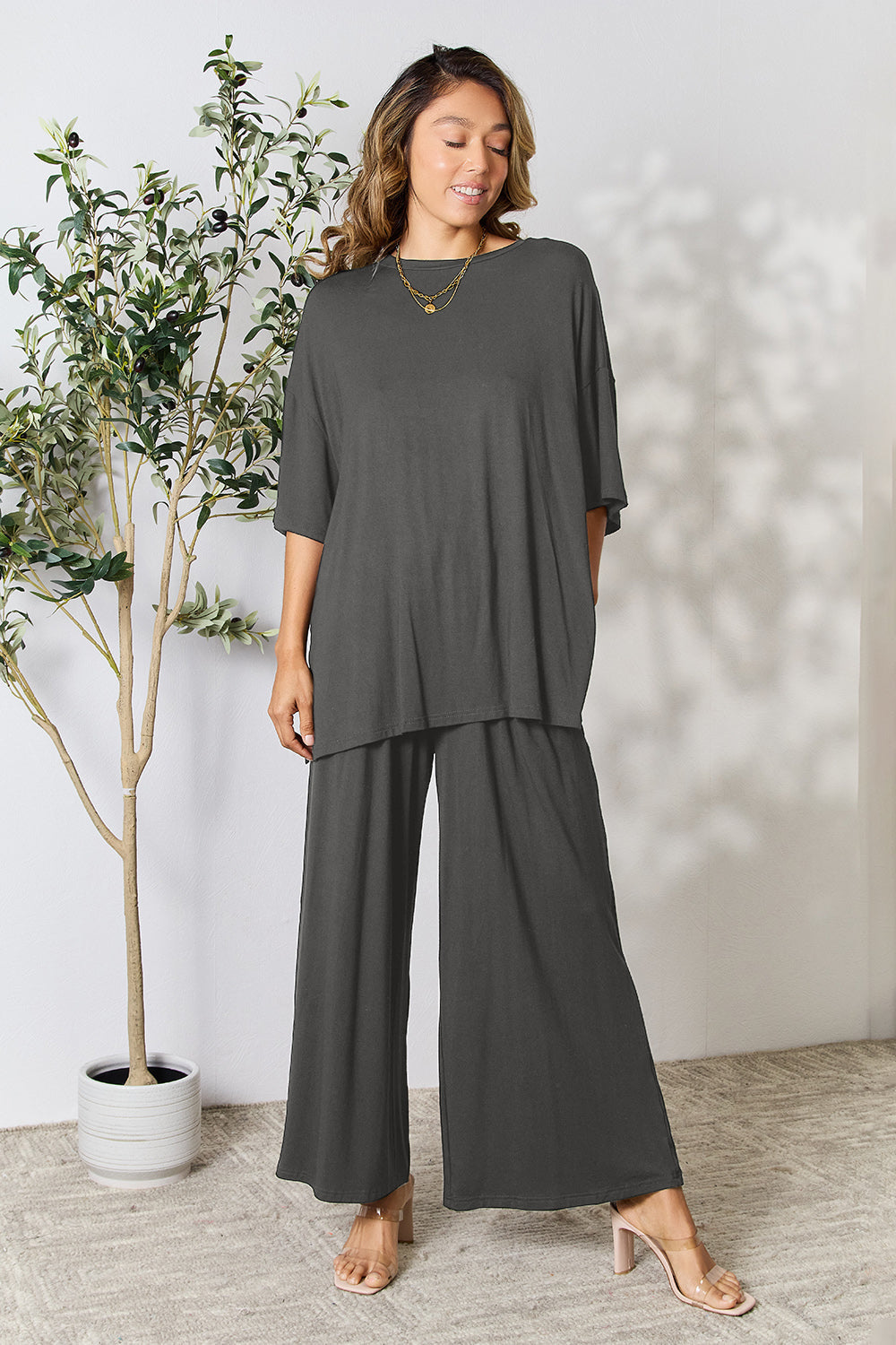 Woman wearing Double Take Round Neck Slit Top and Pants Set in grey, standing by a potted plant, showcasing trendy two-piece outfit.