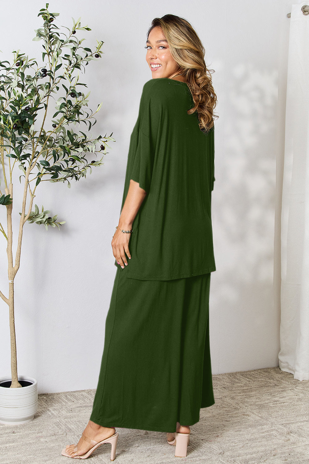 Woman wearing green round neck slit top and pants set, standing next to a plant, showing back view.