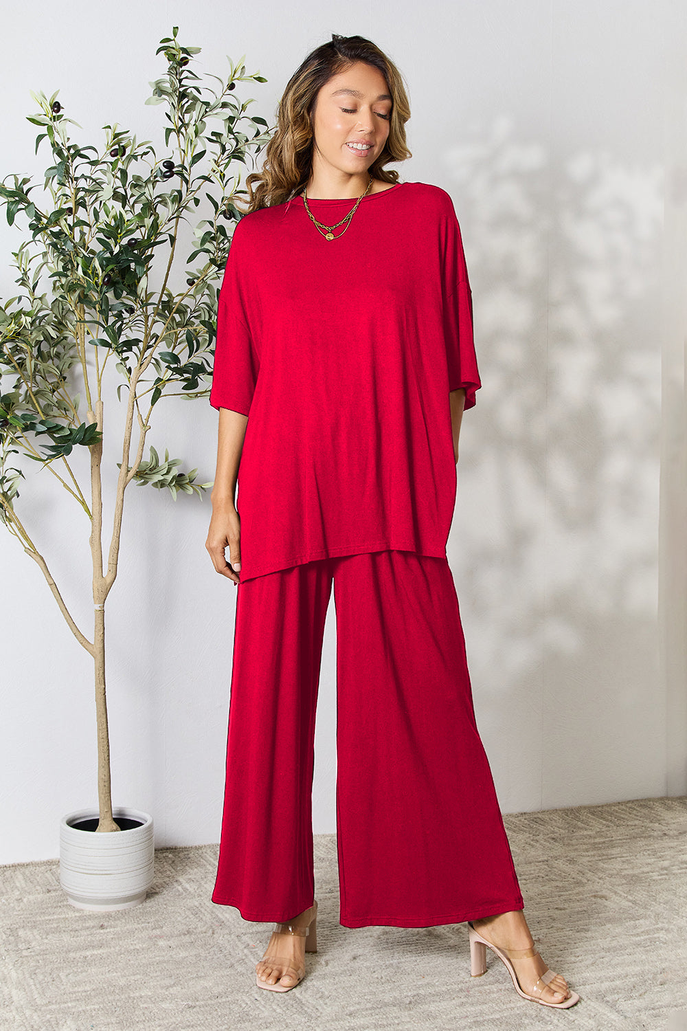 Woman wearing red round neck slit top and pants set, standing near a potted plant.