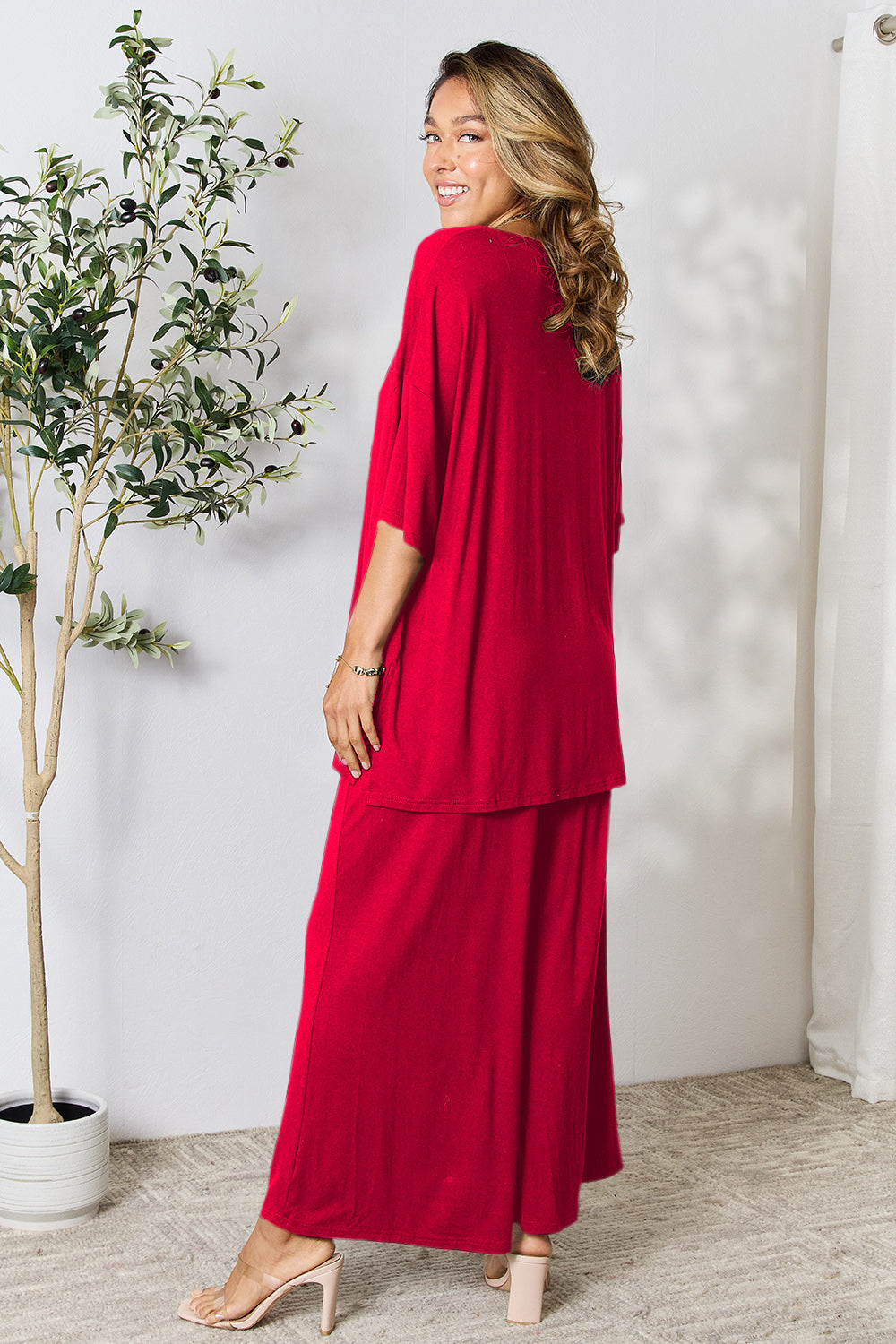 Woman wearing a red double take full-size round neck slit top and pants set, standing in a room with light décor.