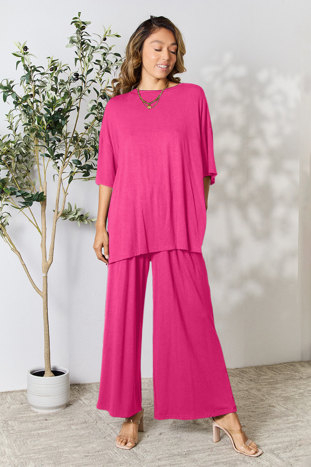 Woman wearing pink two-piece round neck slit top and pants set, standing next to a plant in stylish, comfortable attire.