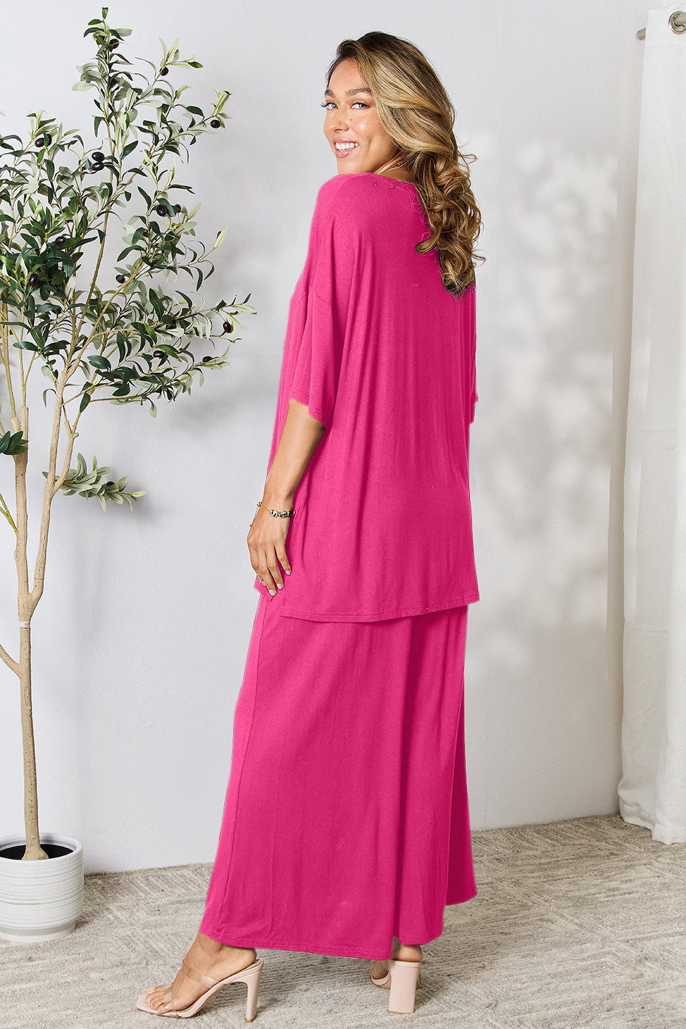 Woman wearing pink round neck slit top and pants set, standing by a plant, showcasing stylish and comfortable fashion outfit.