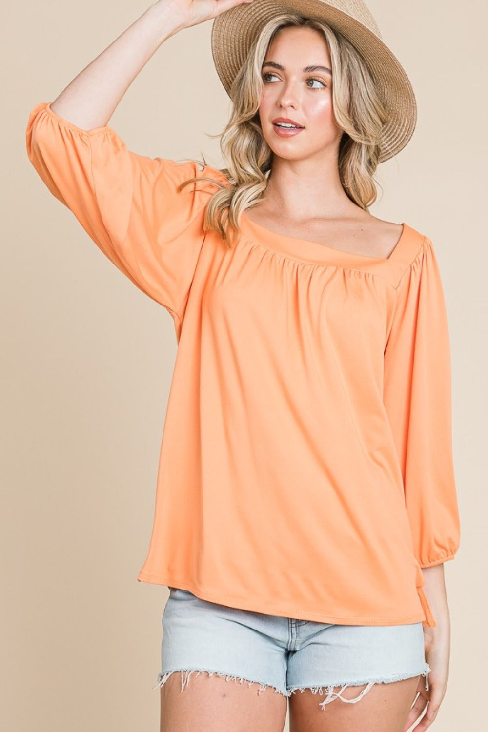 Woman wearing orange square neck puff sleeve top with denim shorts and a hat against a beige background.