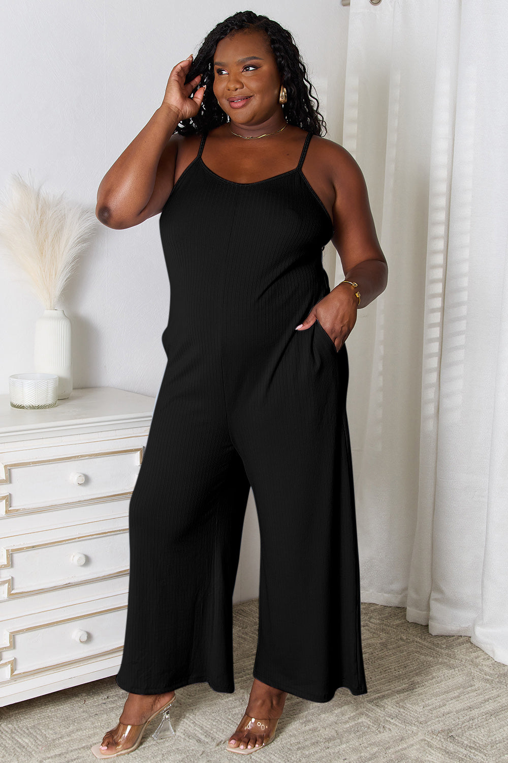 Woman wearing a black spaghetti strap V-neck jumpsuit, standing confidently indoors with a hand on her hip.