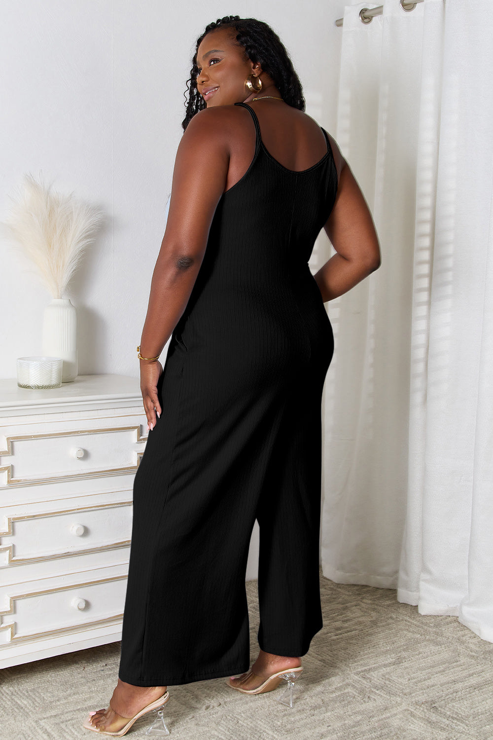 Woman wearing black spaghetti strap V-neck jumpsuit stands in stylish home interior.