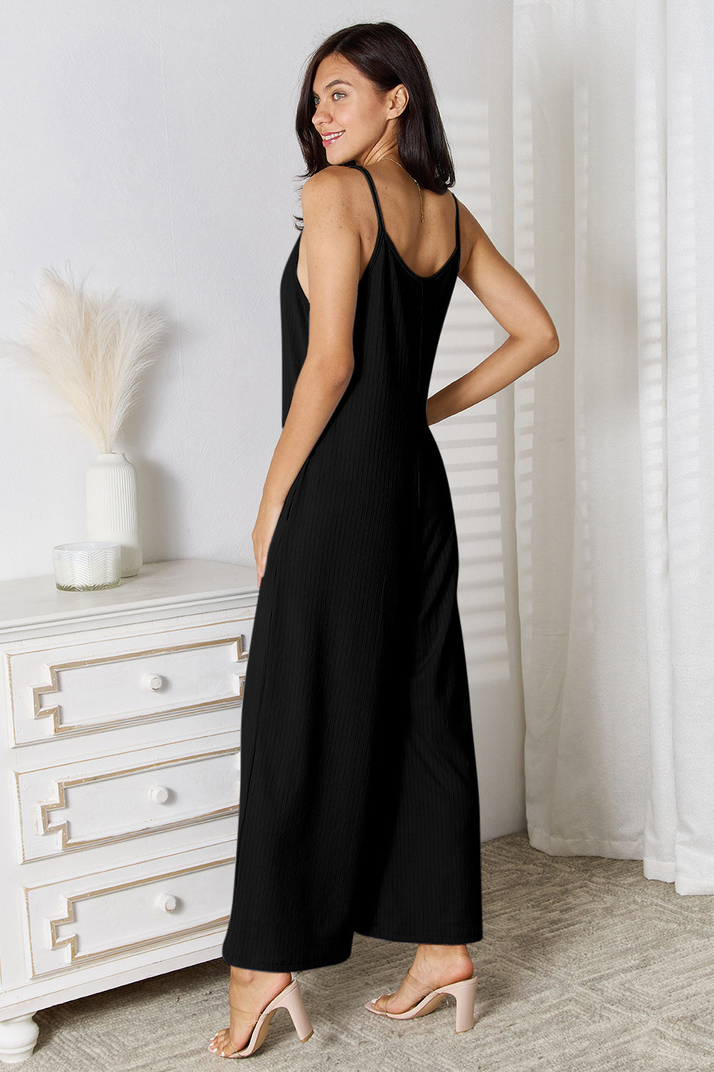 Woman wearing a black spaghetti strap V-neck jumpsuit, standing with her back to the camera in a stylish room setting.