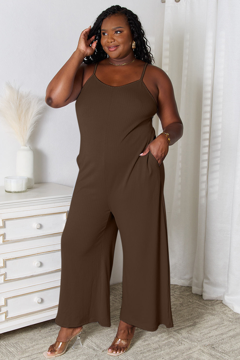 Woman wearing brown spaghetti strap V-neck jumpsuit, standing in chic room with white decor.