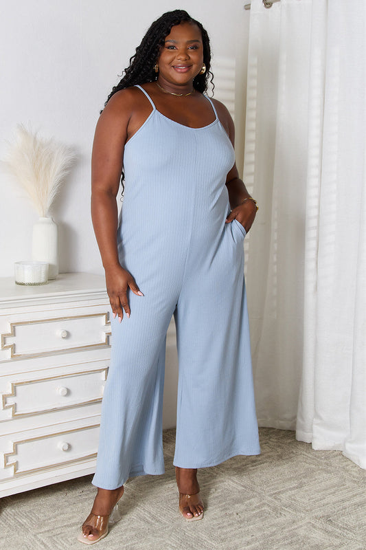 Woman wearing blue V-neck spaghetti strap jumpsuit standing by a dresser in a chic, stylish setting.