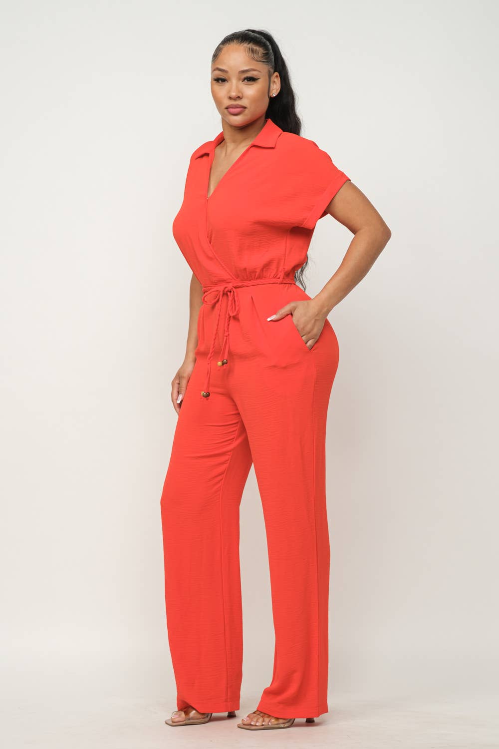 Red waist tie jumpsuit with short sleeves, collared wrap top, and wide leg pants featuring side pockets and elastic shirring.