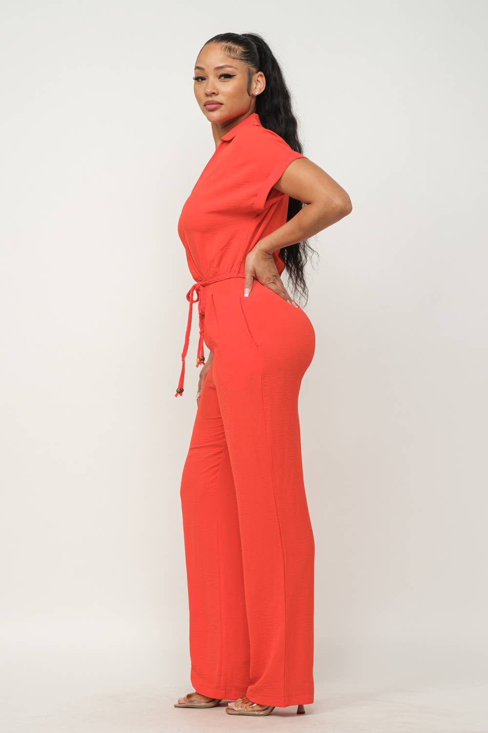 Red open front waist tie jumpsuit with short sleeves, collared wrap top, side pockets, and wide-leg pants, model in profile view.