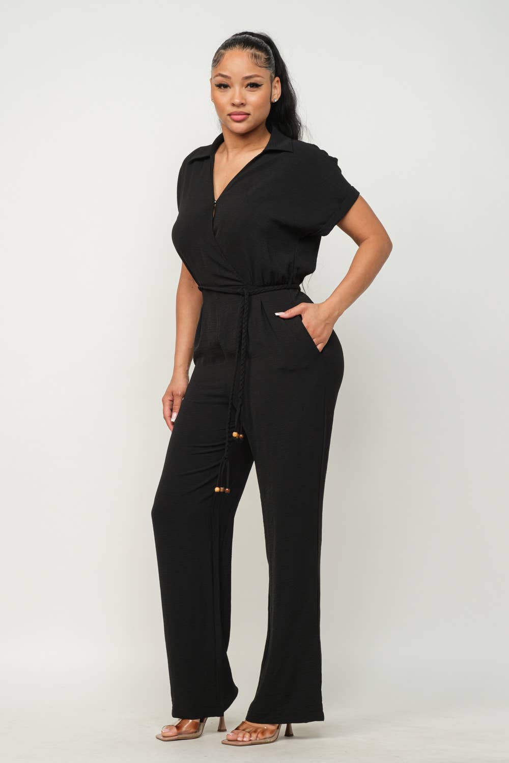 Woman wearing black open front jumpsuit with waist tie and wide leg pants, featuring side pockets and short sleeves.