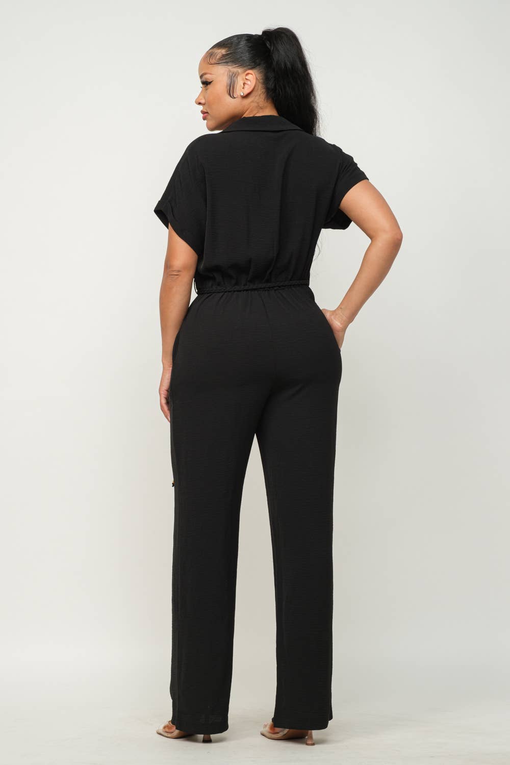 Woman in black open front waist tie jumpsuit with short sleeves and wide leg pants, showcasing back and side pocket details.