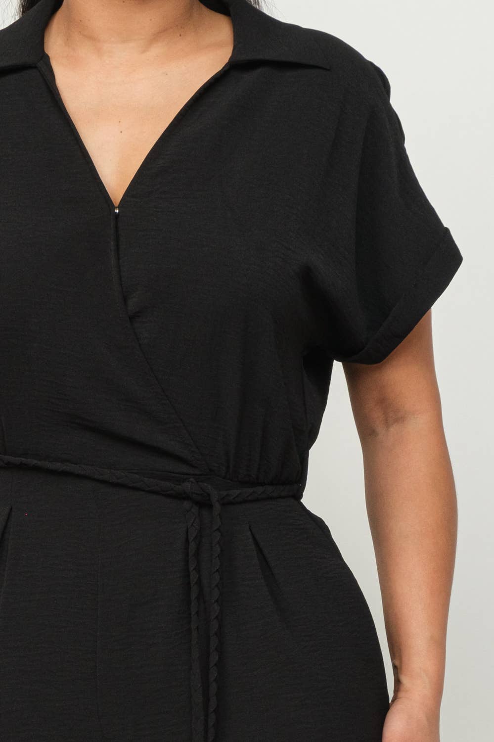 Black wrap top jumpsuit with waist tie and short sleeves, featuring inside button detail and collared neckline.