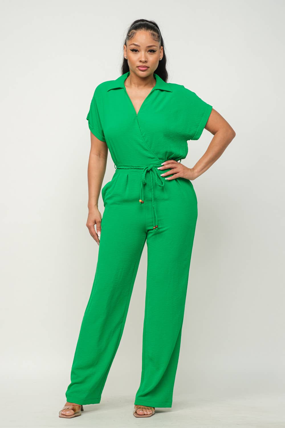 Woman wearing green open front jumpsuit with waist tie, collared short sleeves, and wide-leg pants, styled with beige sandals.