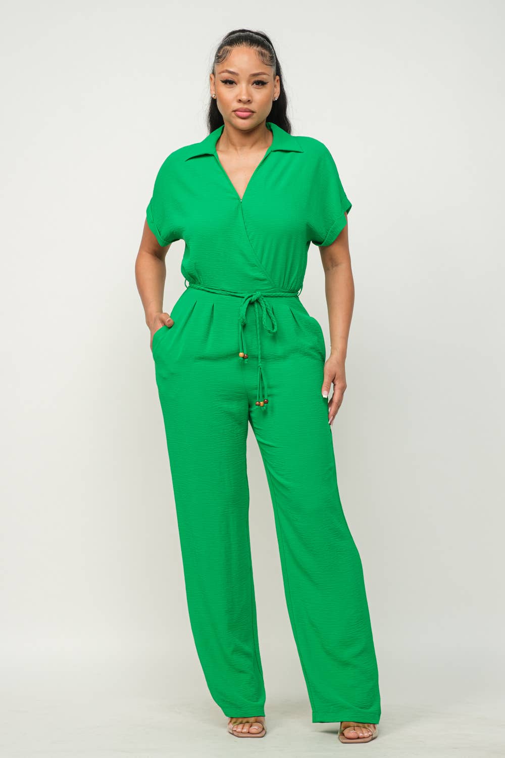 Woman wearing green open front jumpsuit with waist tie, short sleeves, and wide leg pants, showcasing inside button detail and pockets.