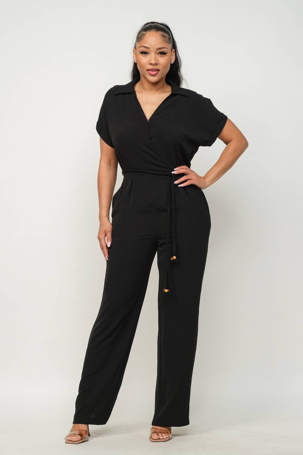 Woman wearing a black open front jumpsuit with waist tie, side pockets, and wide-leg pants. Short sleeve wrap top and elastic shirring.