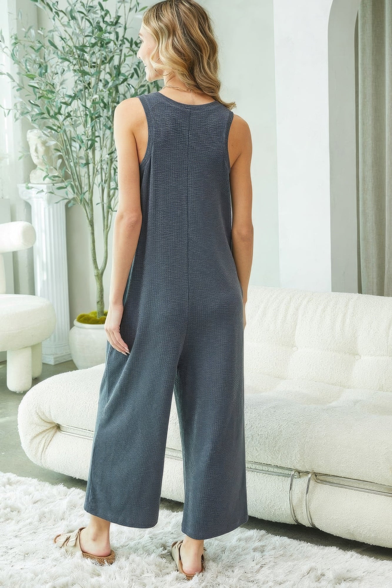 Woman wearing sleeveless charcoal buttondown jumpsuit with a relaxed fit, standing in a cozy living room.