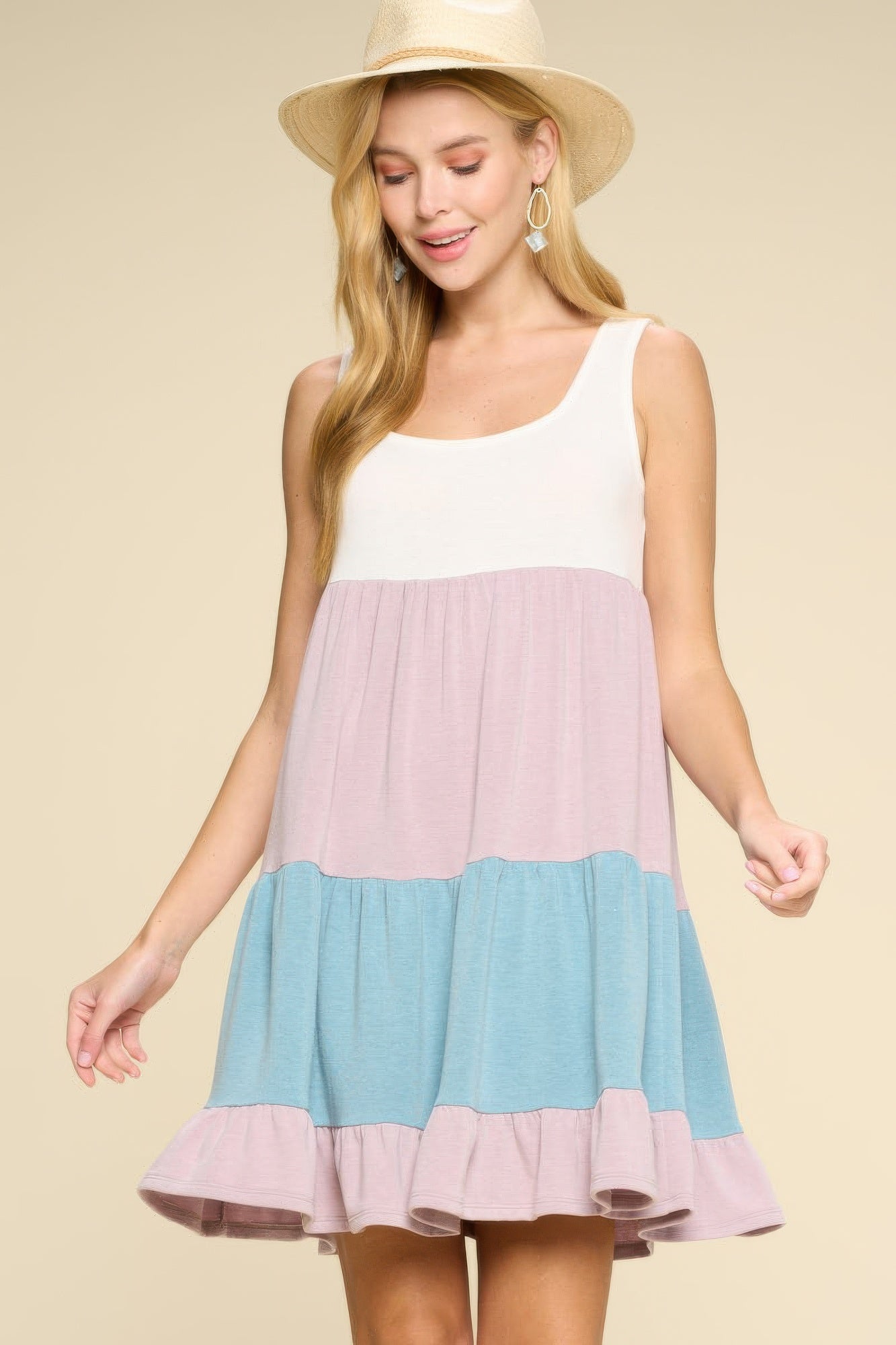 Woman in straw hat wearing sleeveless color-block tiered dress with white, pink, and blue sections, posing against beige background with dangling e...