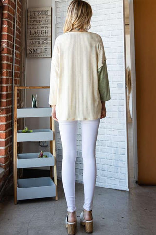 Woman wearing color block round neck dropped shoulder blouse with white leggings, standing in a stylish modern room.