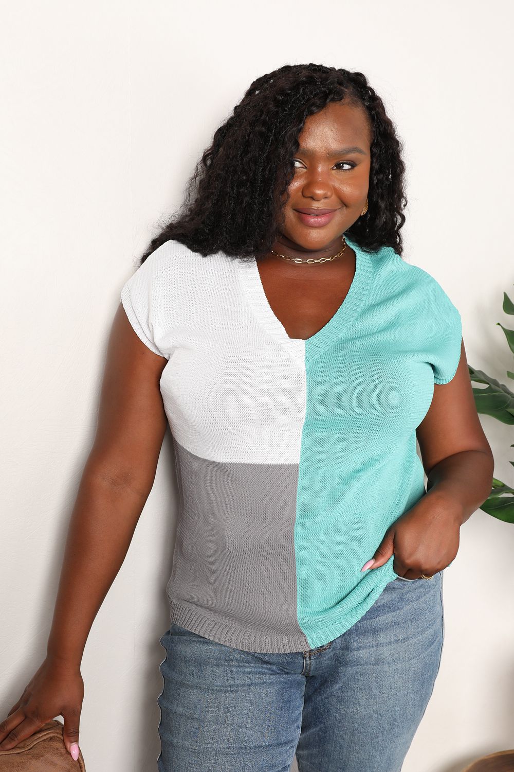 Woman wearing a color block V-neck knit top with short sleeves, featuring white, gray, and teal sections, paired with jeans.