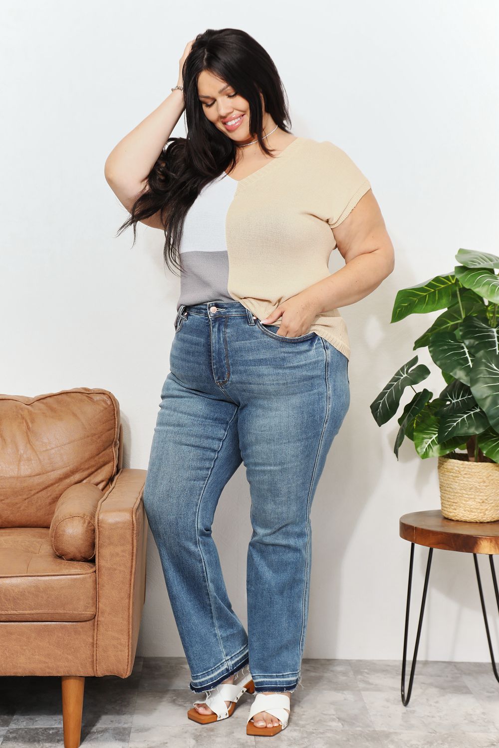 Woman wearing color block V-neck knit top with jeans, standing in a stylish room with plants and a leather chair.