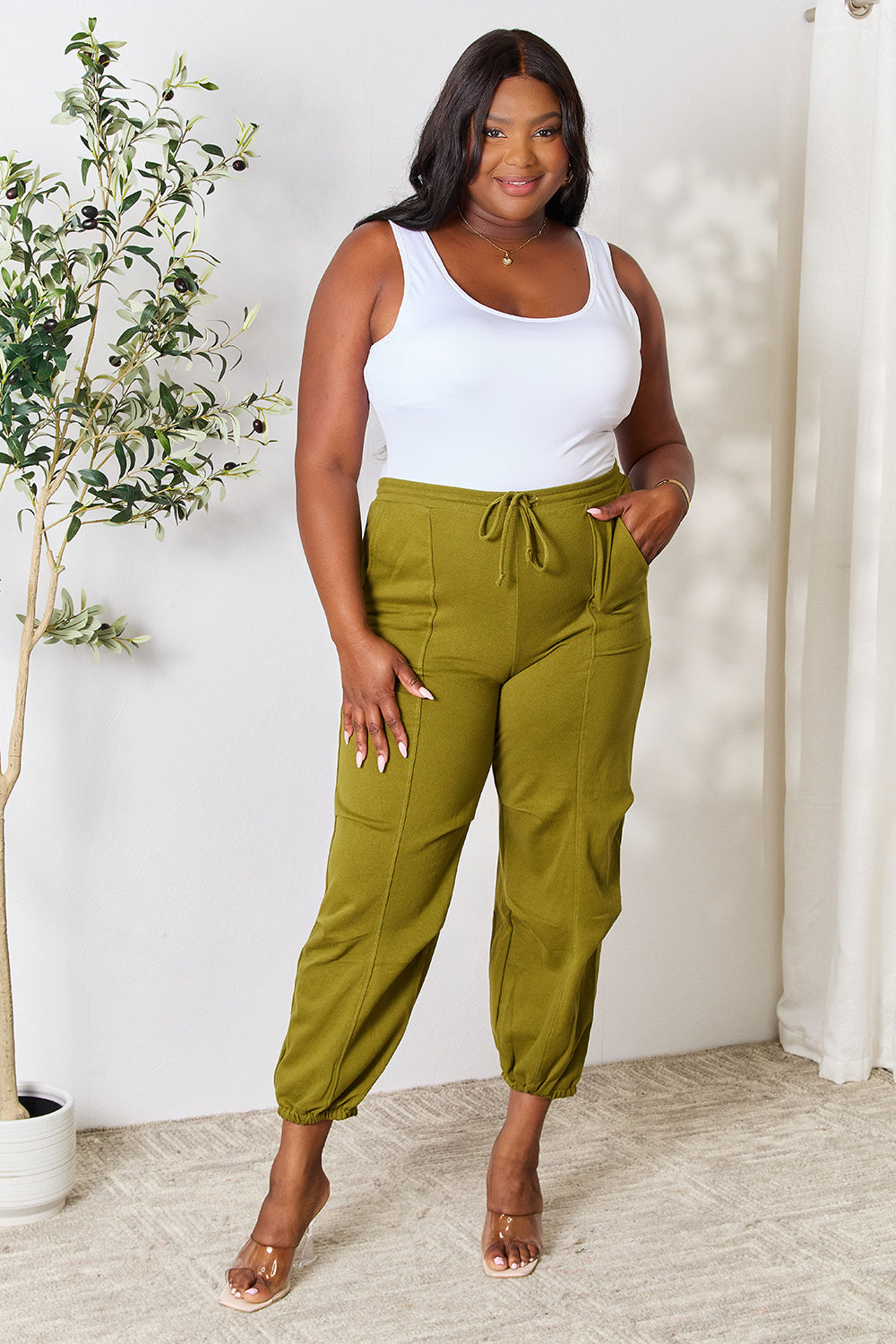 Woman wearing olive green drawstring sweatpants with pockets and a white top, standing in a room with trendy decor.