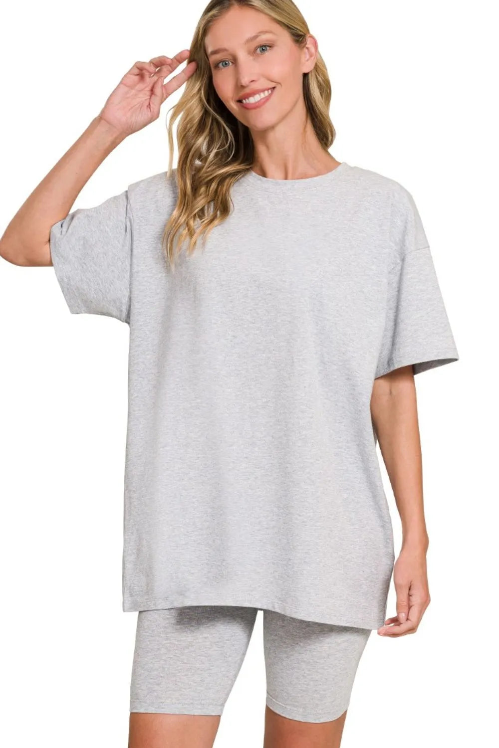 Woman in light gray oversized t-shirt and matching shorts, smiling with hand in her hair, against white background