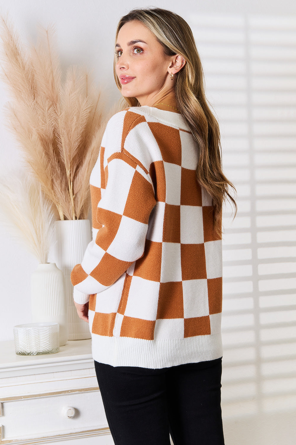 Woman wearing checkered button-up cardigan with dropped shoulder design, standing in a stylish room setting.