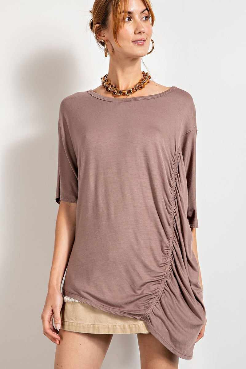 Woman wearing a mocha loose fit top with ruched side detail and boat neckline.