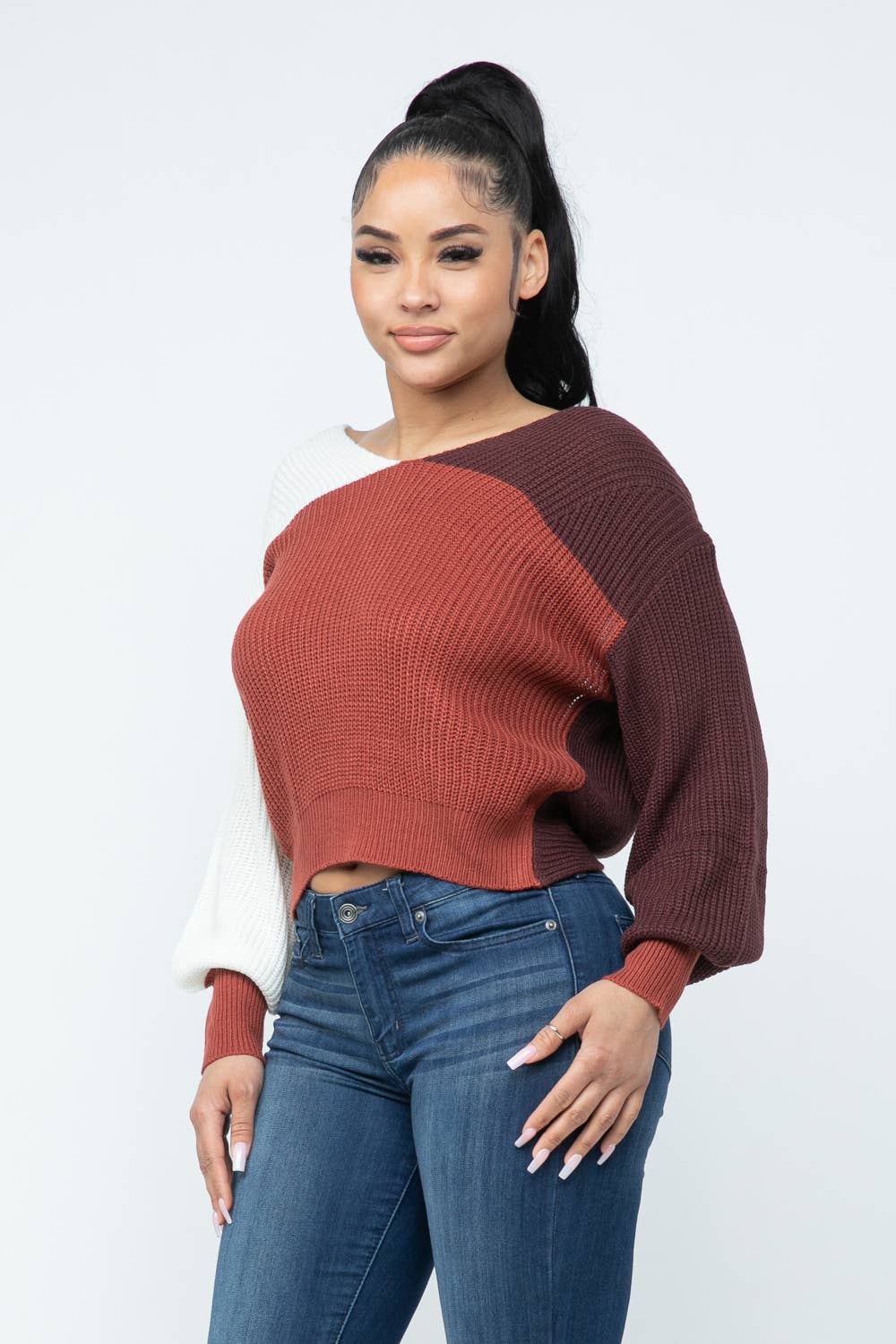 Woman wearing a cozy color block twisted open back sweater in red, white, and brown with a loose fit, paired with blue jeans.