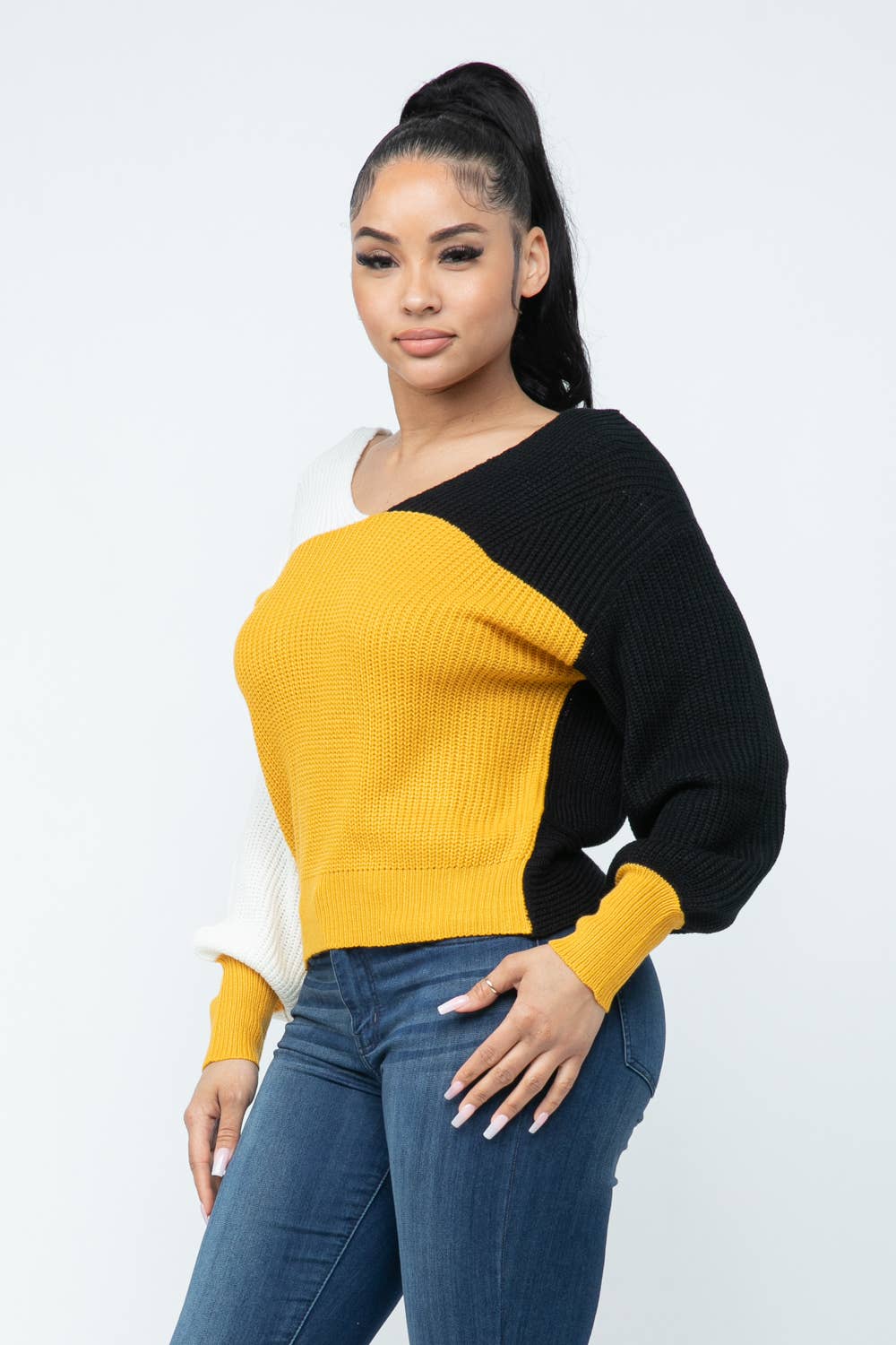 Woman wearing a color block twisted open back loose fit cozy sweater in yellow, black, and white with casual jeans.