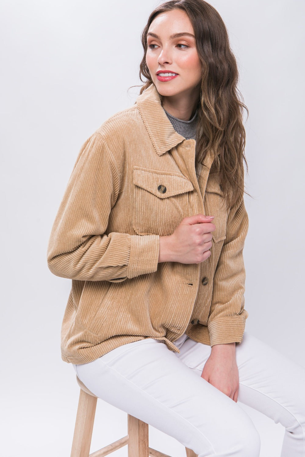 Woman wearing a beige Love Tree corduroy button-up jacket with sherpa lining, sitting on a stool, styled with white pants.