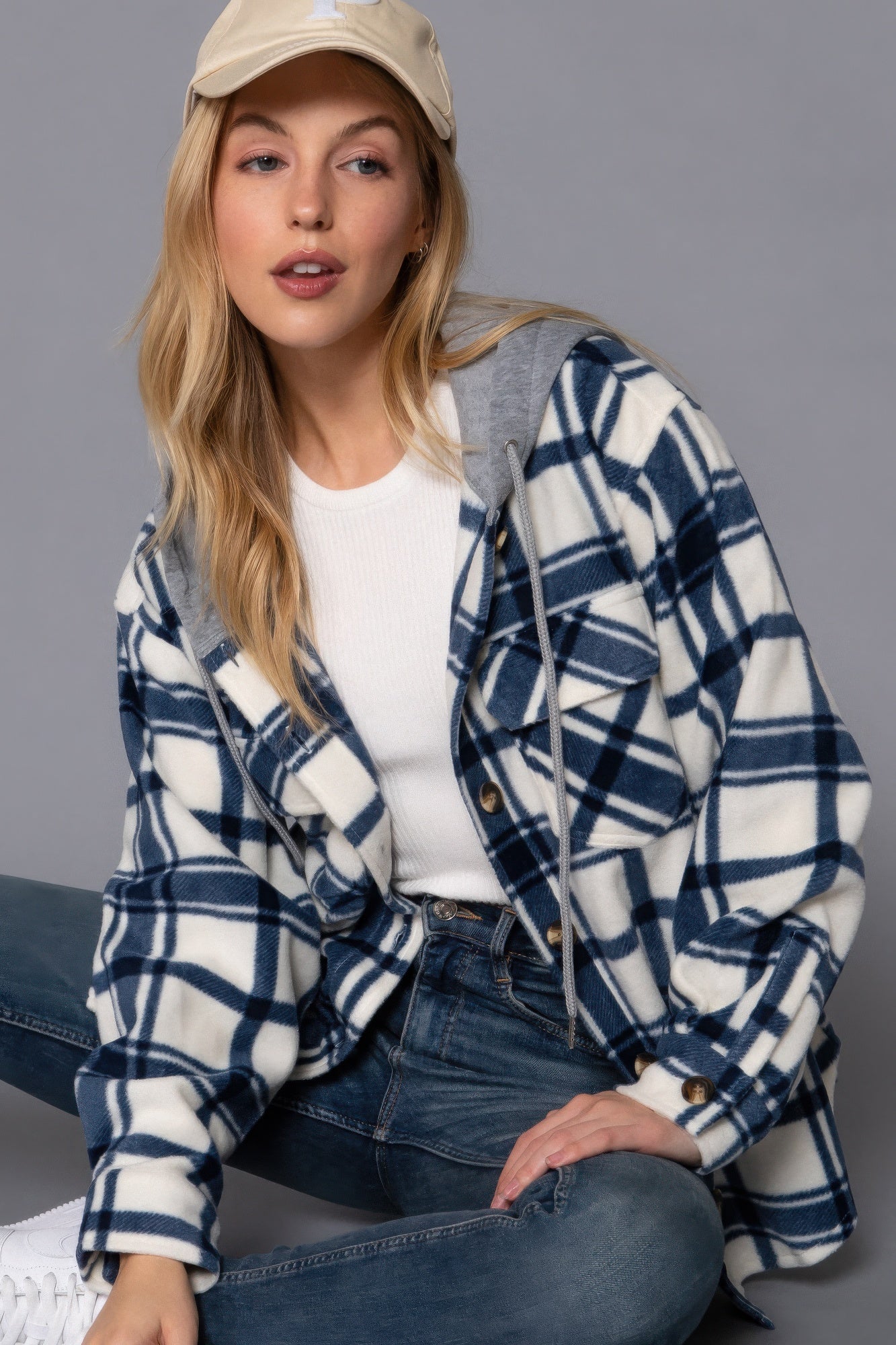 Woman wearing ivory/navy plaid print hoodie fleece jacket styled with a beige cap and blue jeans, showcasing casual chic fashion.