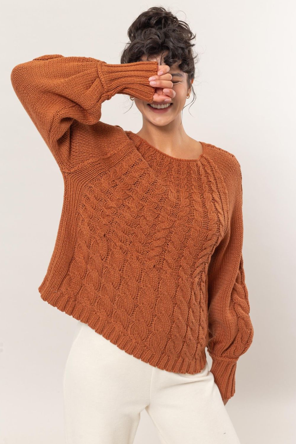 Woman wearing a brown HYFVE cable-knit round neck raglan sleeve sweater with white pants, showcasing cozy winter style.