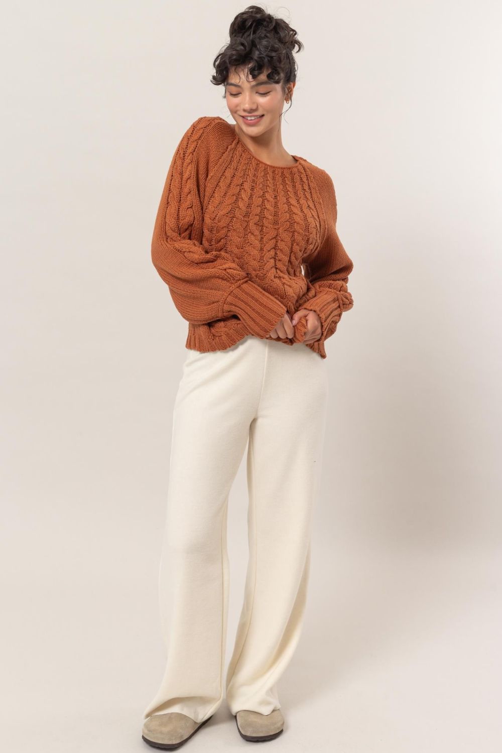 Woman in brown cable-knit round neck raglan sleeve sweater with white pants, showcasing cozy and chic winter fashion.