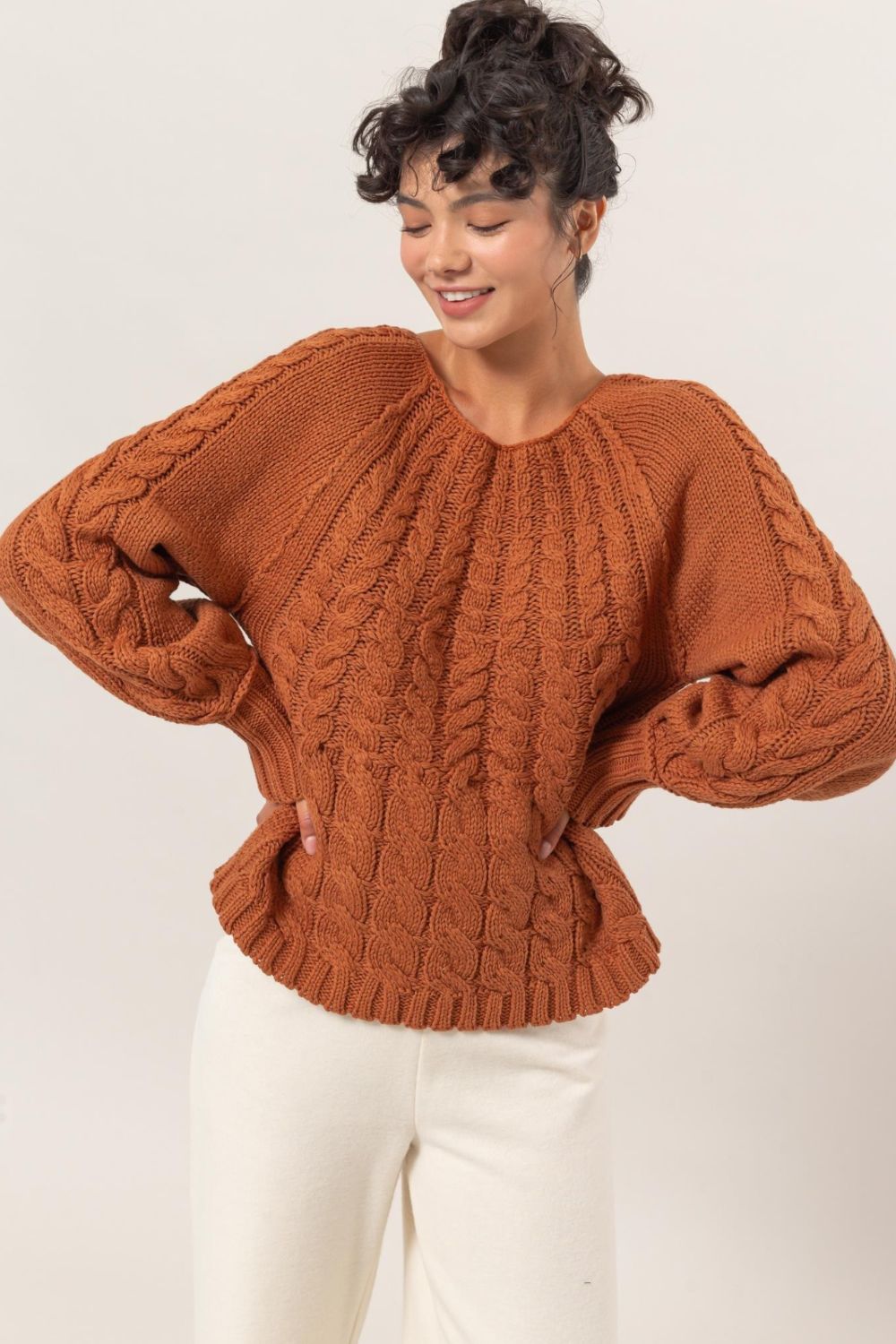 Woman wearing HYFVE cable-knit round neck raglan sleeve sweater in cozy brown, paired with white pants. Perfect winter fashion staple.