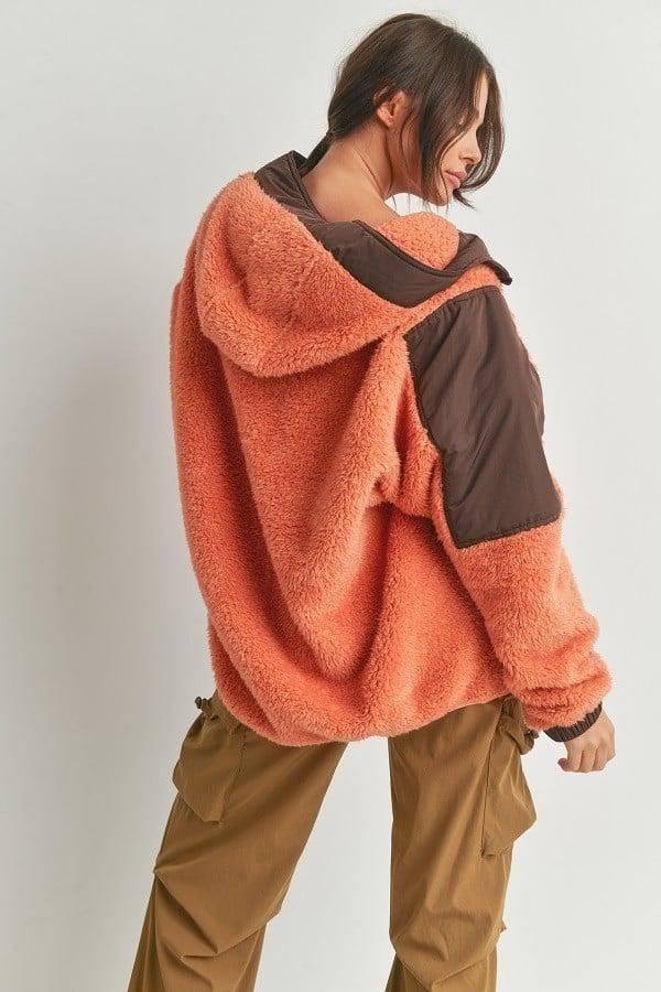 Woman wearing two-toned cozy hooded sweater in orange and brown, showcasing back design. 100% polyester, perfect for fall fashion.