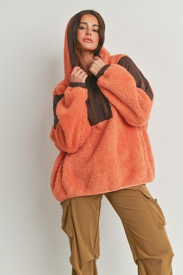 Two-toned cozy hooded sweater in orange and brown, made of 100% polyester, shown on a model against a light background.